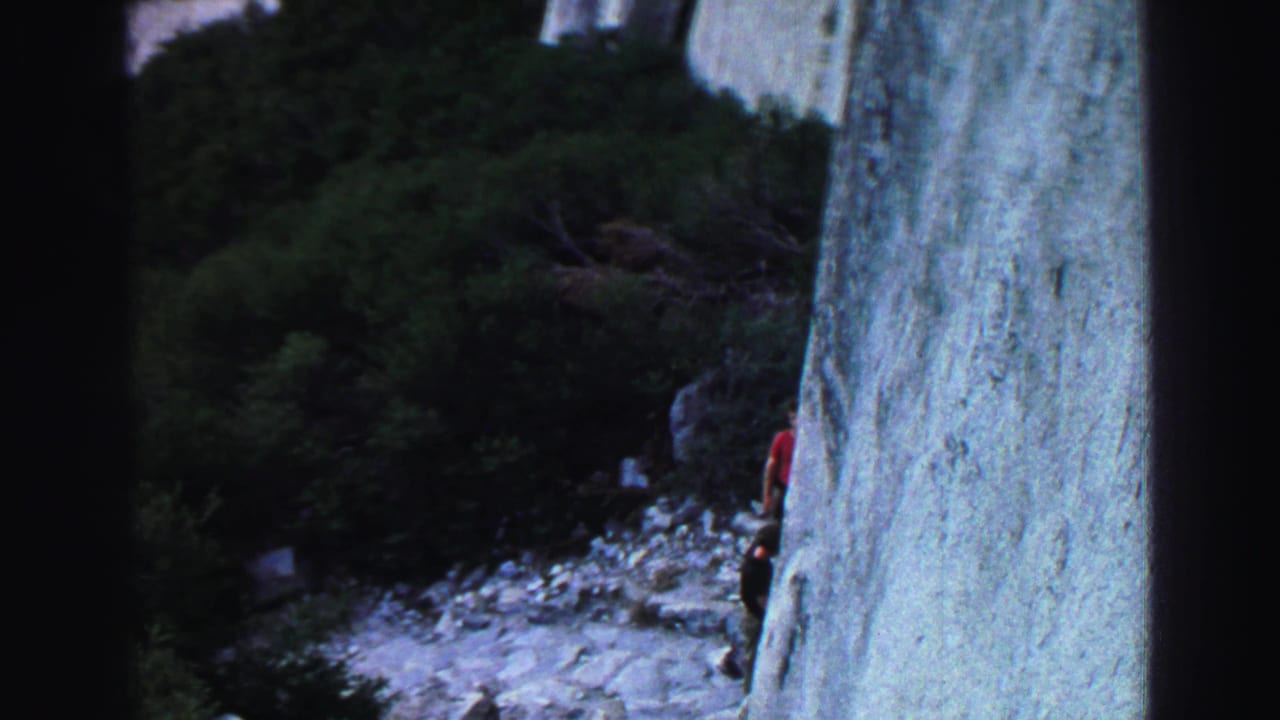 Distant climber beside a pale granite wall with shrubs and rocks below.
