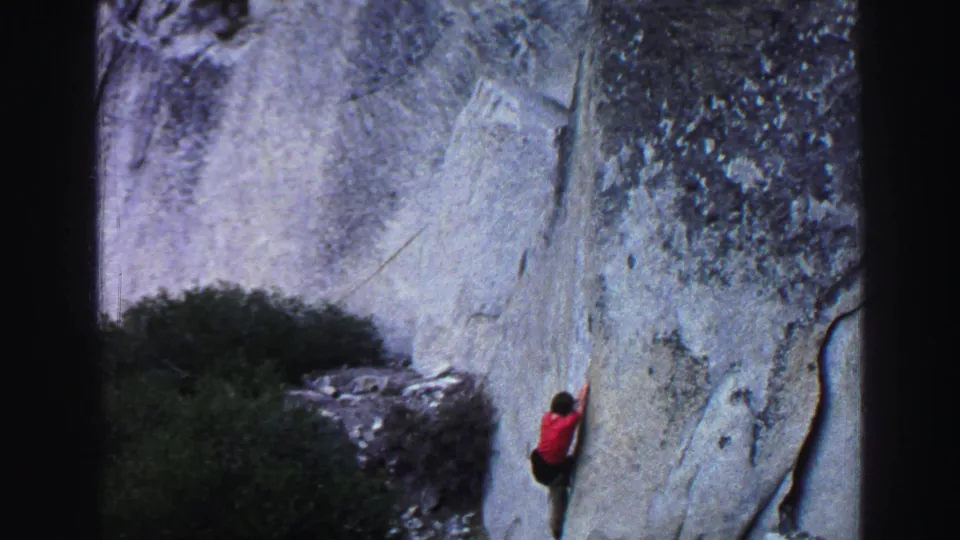 1969 Lake Tahoe 8mm frame of a red-shirted climber using handholds on a steep granite face above brush and talus.