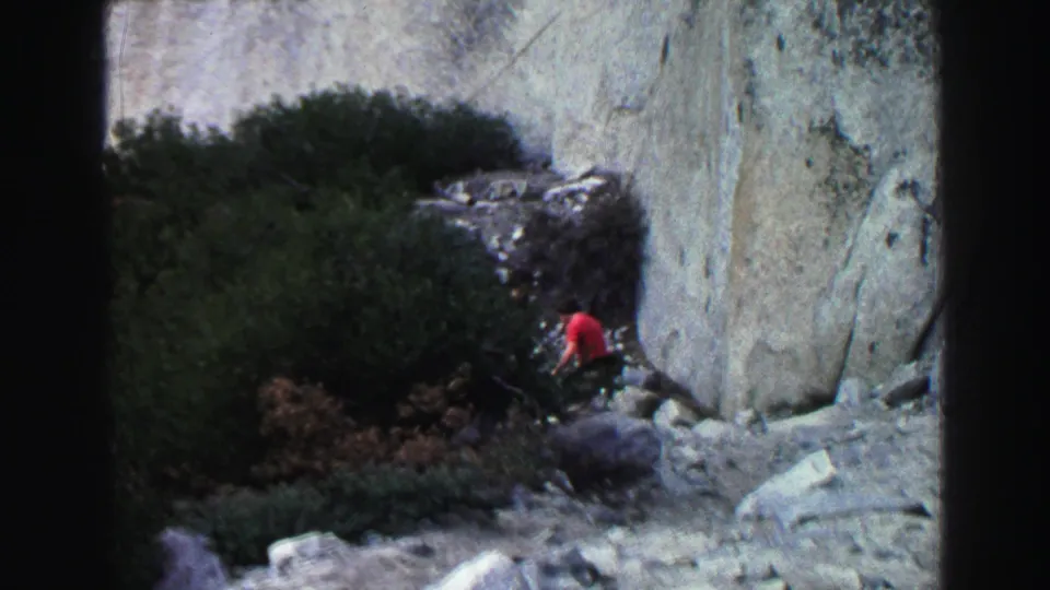 1969 Lake Tahoe home-movie frame showing a distant person in red near shrubs and a pale granite rock face.