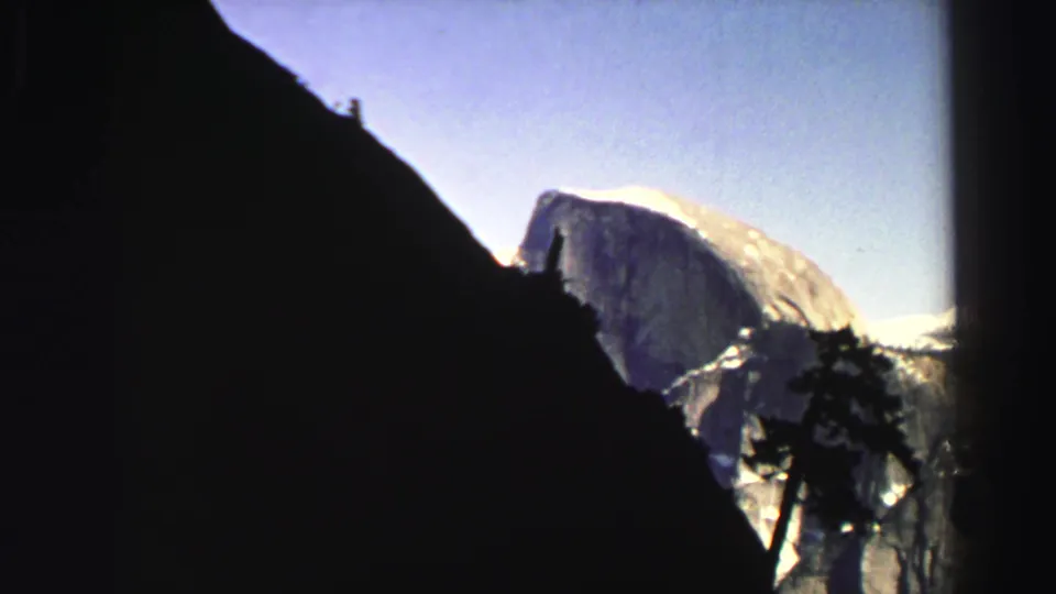 1969 mountain home-movie frame with a tiny ridge figure silhouetted against sky and a bright rock formation in the distance.