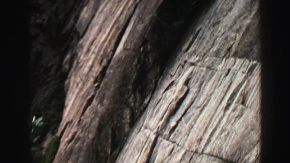 Close 8mm frame of a steep, textured rock face with vertical striations, deep shadows, and small plants near the lower edge.