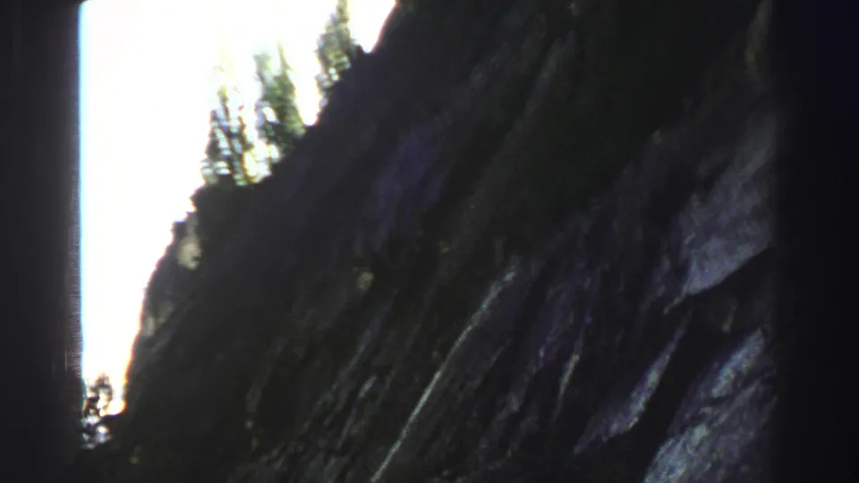 Handheld 1969 Lake Tahoe footage frames a dark cliff wall beside bright sky and small evergreen silhouettes.