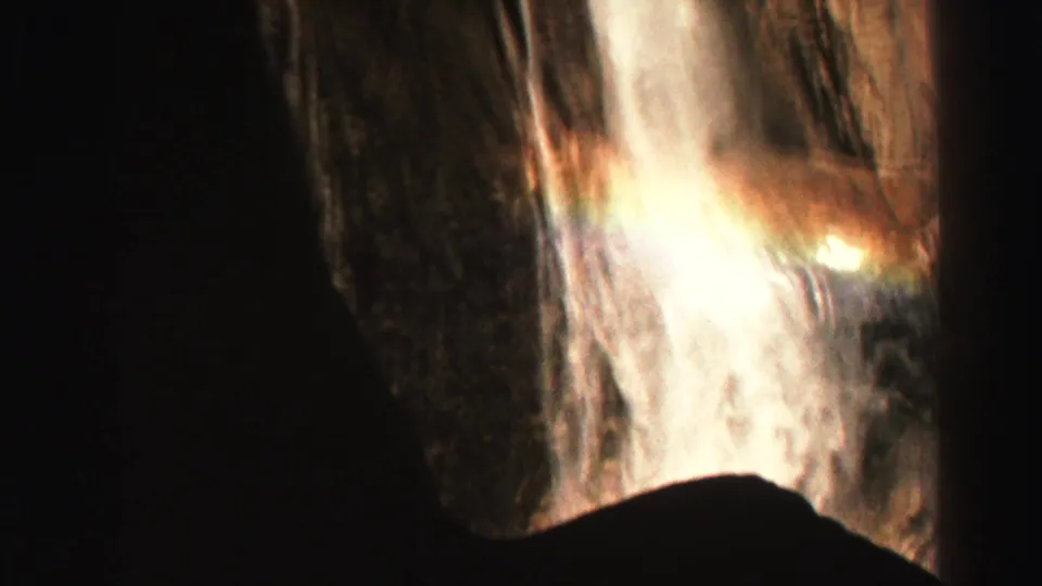 1969 8mm Lake Tahoe area footage frame with waterfall spray, warm glare, and rainbow-like flare against shadowed canyon rock.