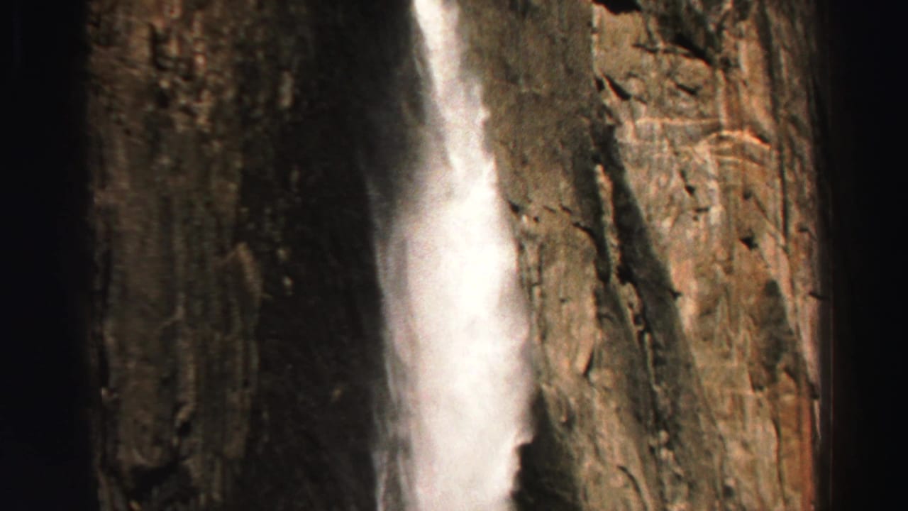 Waterfall dropping beside a rugged rock wall in a dark canyon-like setting.