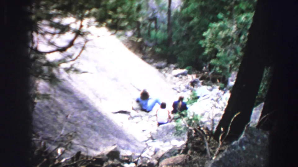 1969 Lake Tahoe home-movie frame showing three people resting on a sunlit rocky slope beneath conifer branches.