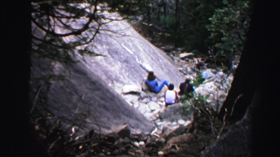 1969 Lake Tahoe frame showing three distant people seated beside a smooth rocky slope within a shaded forest setting.