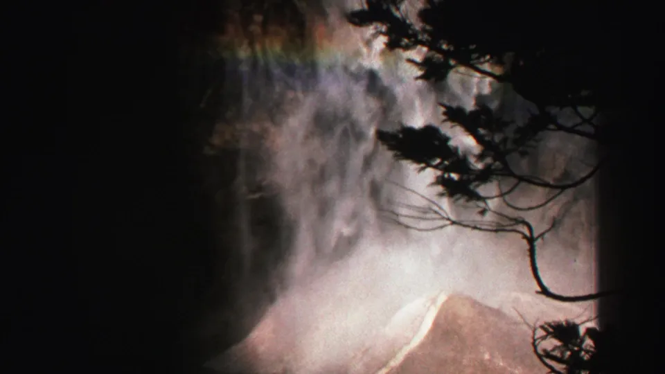1969 Lake Tahoe 8mm frame of waterfall mist, a faint rainbow, and dark branch silhouettes near the cliff base.