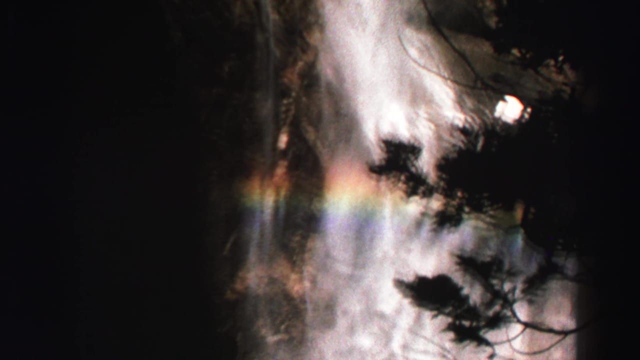 Bright waterfall mist behind dark tree silhouettes with a faint rainbow band.