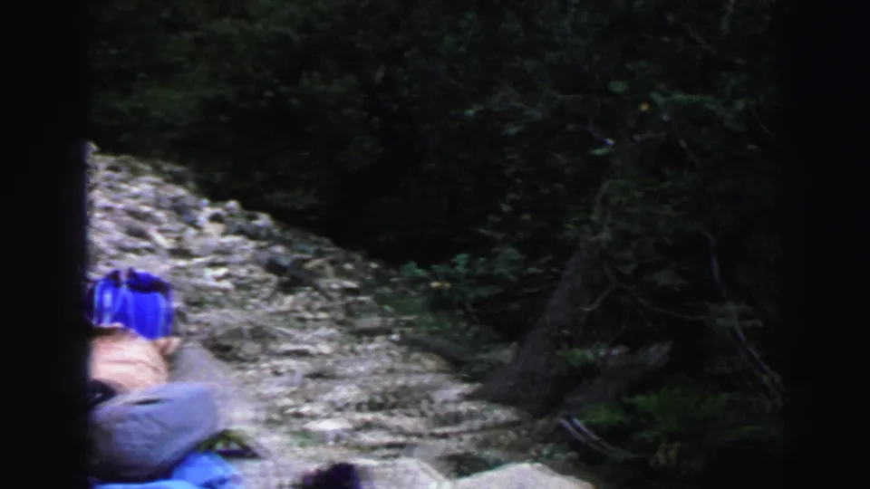Handheld 1969 Lake Tahoe trail shot showing blue gear partly in frame beside a stony path and dense shrubs.