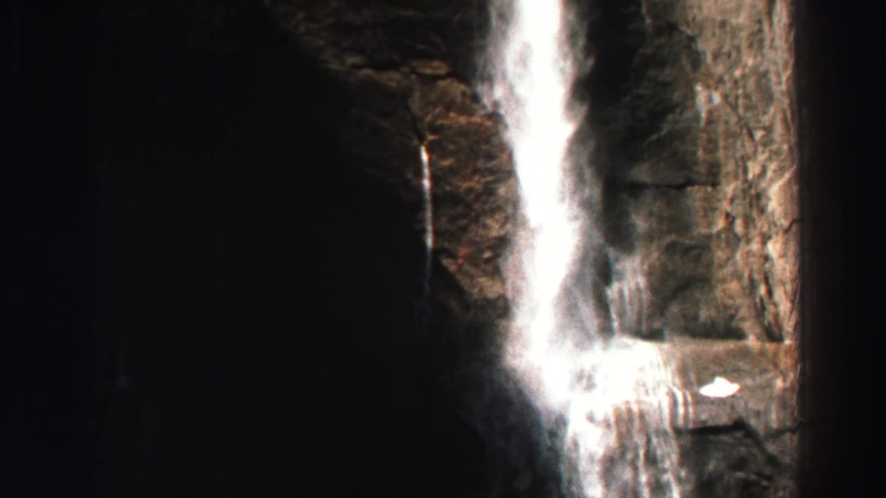 Waterfall descending a rocky cliff in dark, high-contrast forest light.