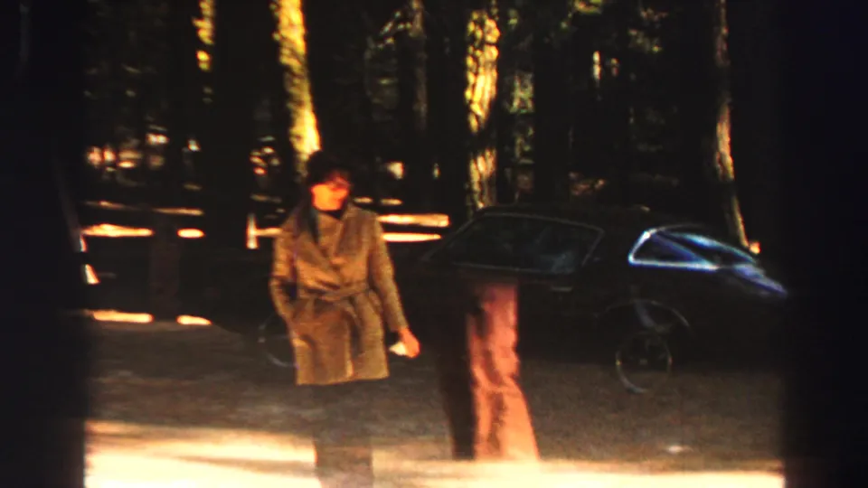 Lake Tahoe home-movie frame with two adults near a dark parked car under tall forest trees.