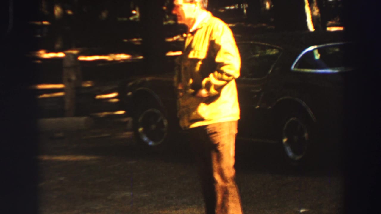 Man in a yellow jacket stands near a dark parked car in a wooded roadside area.