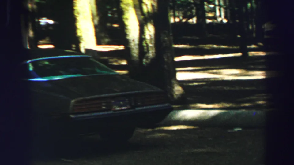 A dark classic car sits beside trees and dappled pavement in Lake Tahoe 1969 home-movie footage.