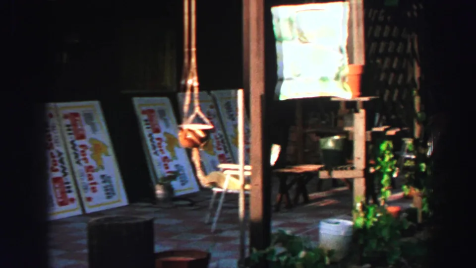 1969 Lake Tahoe 8mm frame showing white sale boards propped in a dim open area with plants, stools and hanging macrame holders.