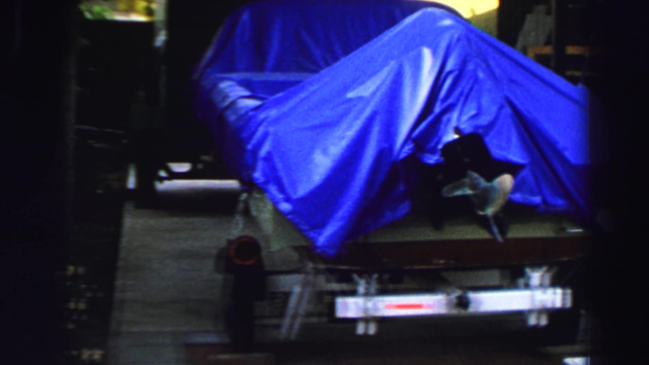 1969 Lake Tahoe 8mm frame showing a boat covered with a vivid blue tarp on a trailer near trees and driveway shade.