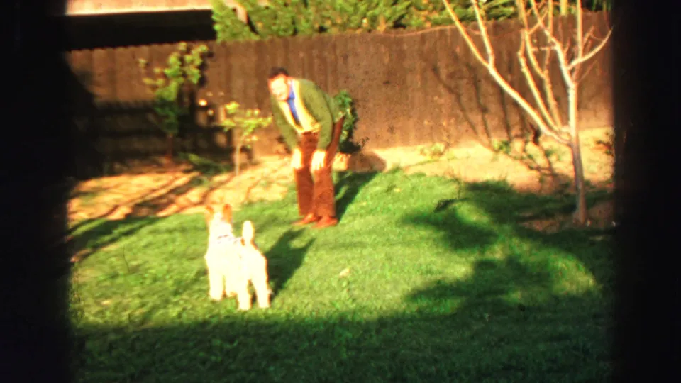1969 Lake Tahoe home-movie frame of an adult man in a green sweater bending toward a small white dog on the lawn.