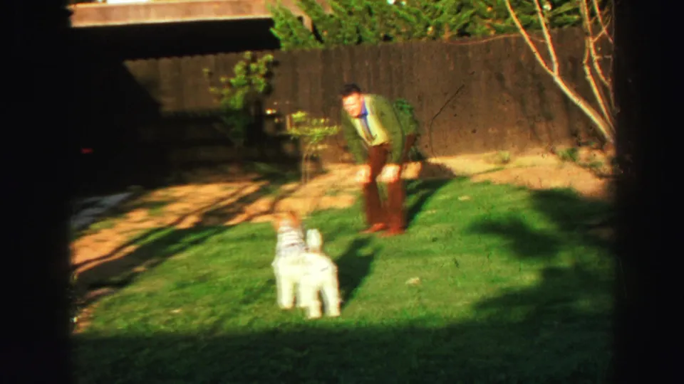 1969 Lake Tahoe home-movie frame of a man in a green jacket bending toward a shaggy white dog in a fenced backyard.