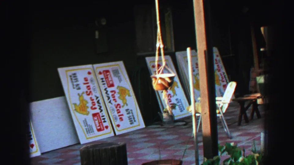 1969 Lake Tahoe 8mm frame showing colorful For Sale signs by a dark exterior wall, with a hanging planter and white patio chair nearby.