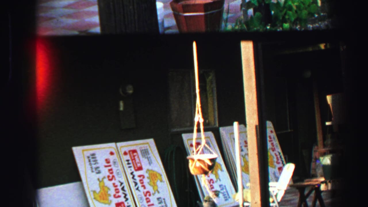 Sale signs, a hanging planter, and patio furniture appear beside a shaded wall.