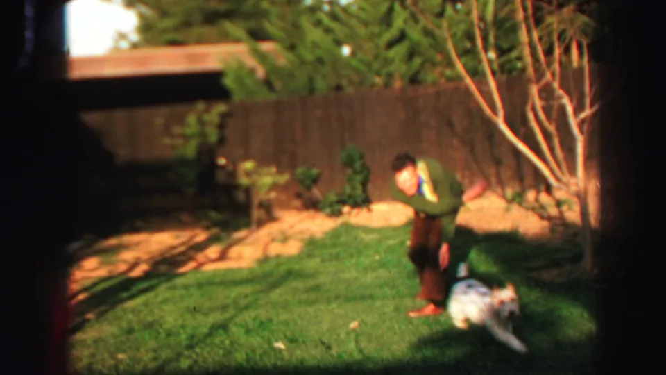 1969 Lake Tahoe backyard footage frame showing a man in casual clothes with a small white dog on a lawn bordered by a wooden fence and trees.