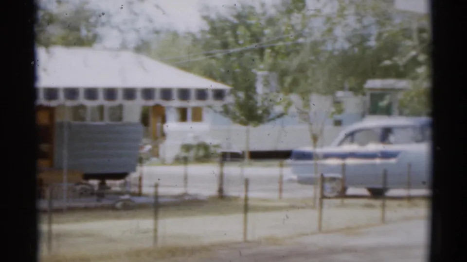 1959 Disneyland California home-movie frame showing a camper trailer, light-colored car, awning, trees, and fence viewed through a window.