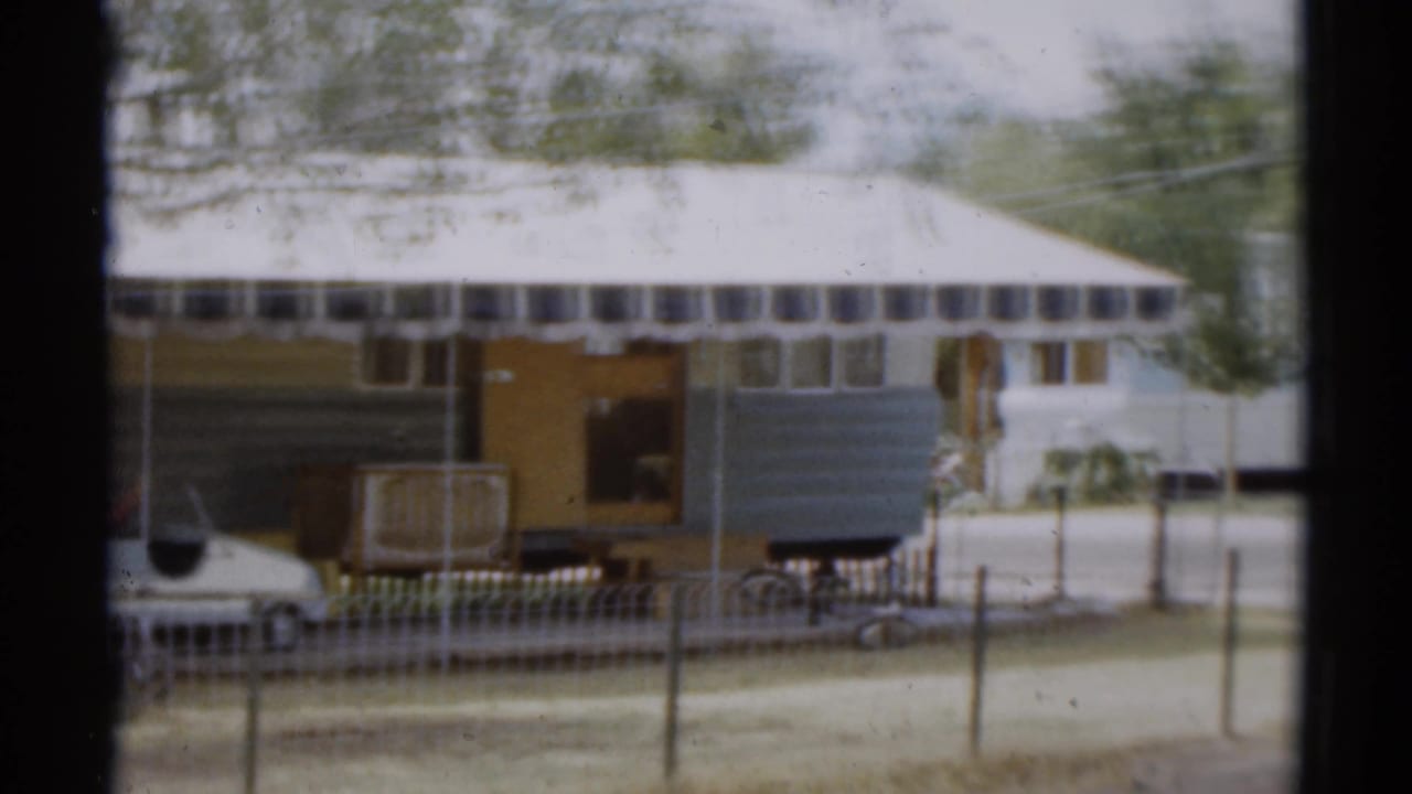 Large camper trailer with open doorway, awning, fence, trees, and a parked car partly visible.
