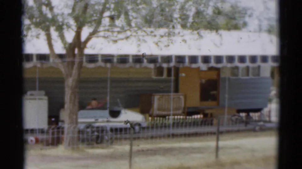 1959 Disneyland California campsite view with trailer, tree, fence, parked car, patio items, and a small visible person near the setup.