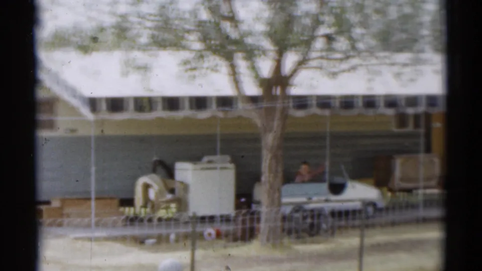 1959 Disneyland California frame showing a trailer campsite with tree, fence, parked car, outdoor furniture, and a small person near the car.