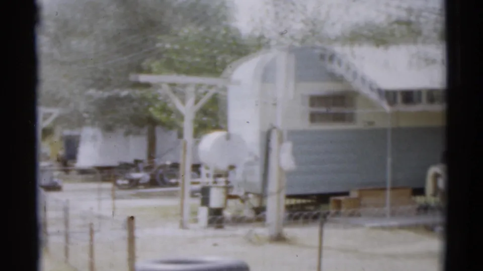 1959 8mm frame showing mobile homes with awnings, trees, fence posts, outdoor equipment, and dark trucks in the Disneyland, California area.