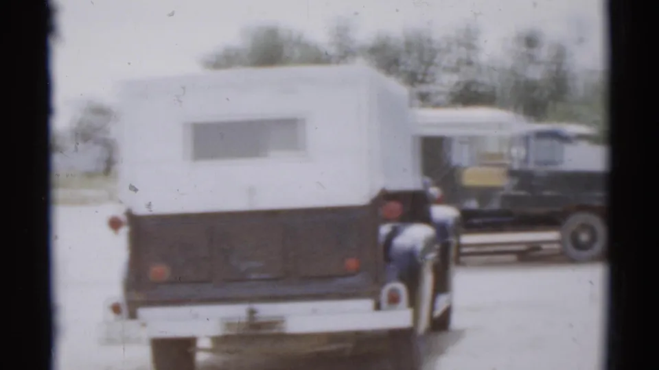 1959 8mm frame of a two-tone trailer or camper from behind, with indistinct people nearby and a dark truck in the background.