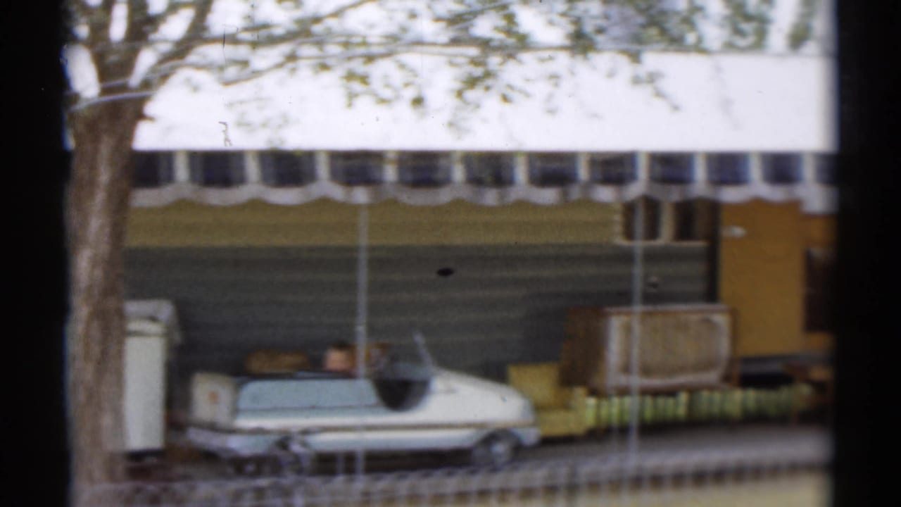 Soft 8mm frame of a Disneyland-area trailer or chalet exterior, striped awning, trees, and a light-colored vintage car.