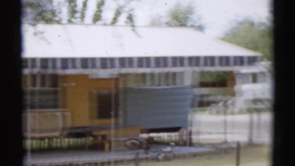 1959 home-movie view of a trailer entrance area with striped awning, fence posts, outdoor furnishings, and a child's pedal car.