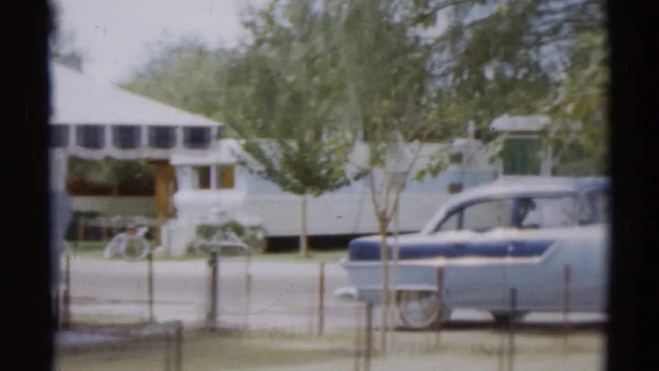 Disneyland-area 1959 frame showing a trailer exterior, small tree, bicycles, fence posts, and a blue-and-white vintage car.