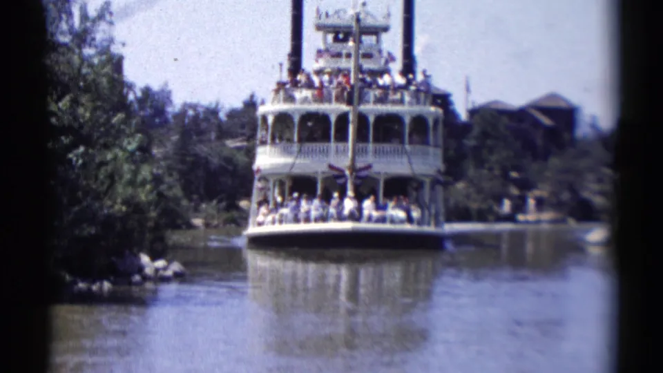 1959 Disneyland riverboat ride seen from a visitor viewpoint, with a crowded white boat moving along calm water between trees.