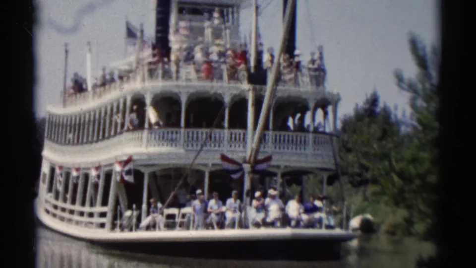 White multi-deck riverboat at Disneyland, with visitors in summer clothing visible from a handheld 8mm view.