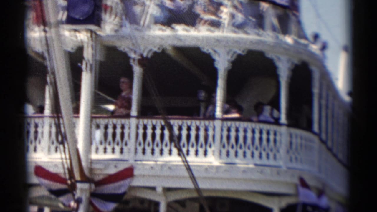 Side view of the Disneyland riverboat, showing curved white railings, shaded passengers, support poles, and bunting on the deck edge.