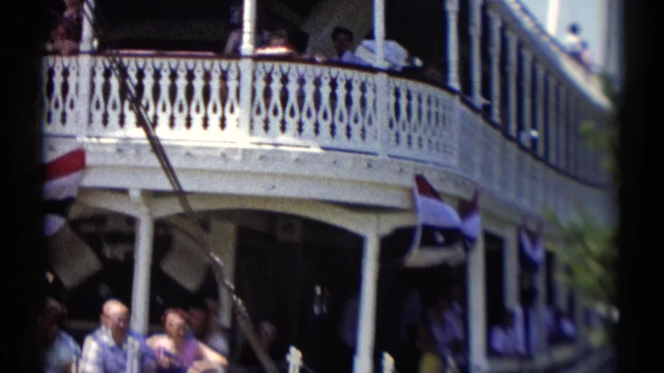 Disneyland riverboat decks pass close to camera, with ornate white railings, shaded cabins, standing passengers, and bunting.