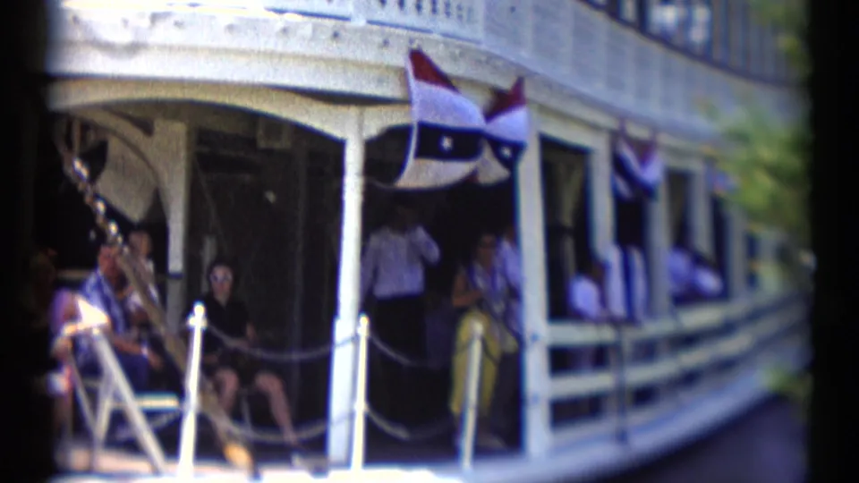 1959 Disneyland riverboat side deck with passengers near a dark doorway, white posts, rope rails, and patriotic bunting.