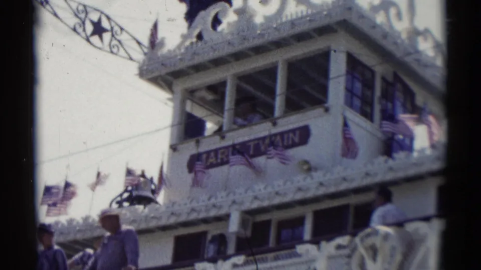 1959 Disneyland frame showing the Mark Twain Riverboat facade, American flags, passengers, windows, and decorative white trim.