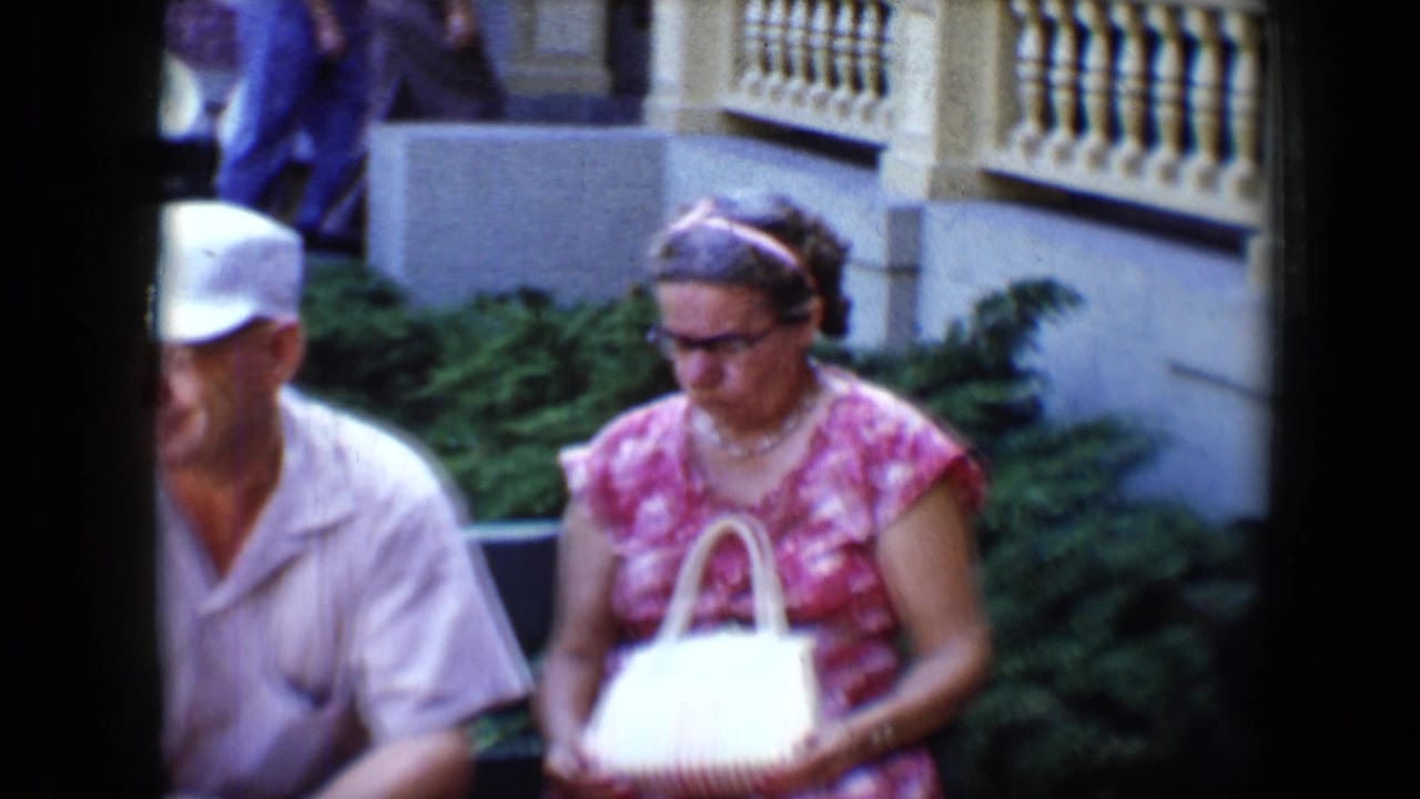 Woman in pink floral dress with white handbag sits beside man in white cap outdoors.