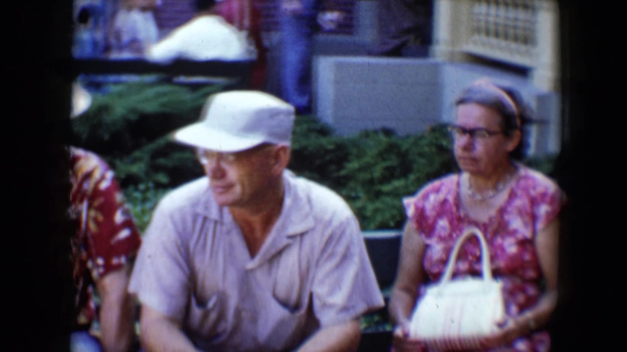 Man in white cap and woman with handbag sit side by side near park landscaping.