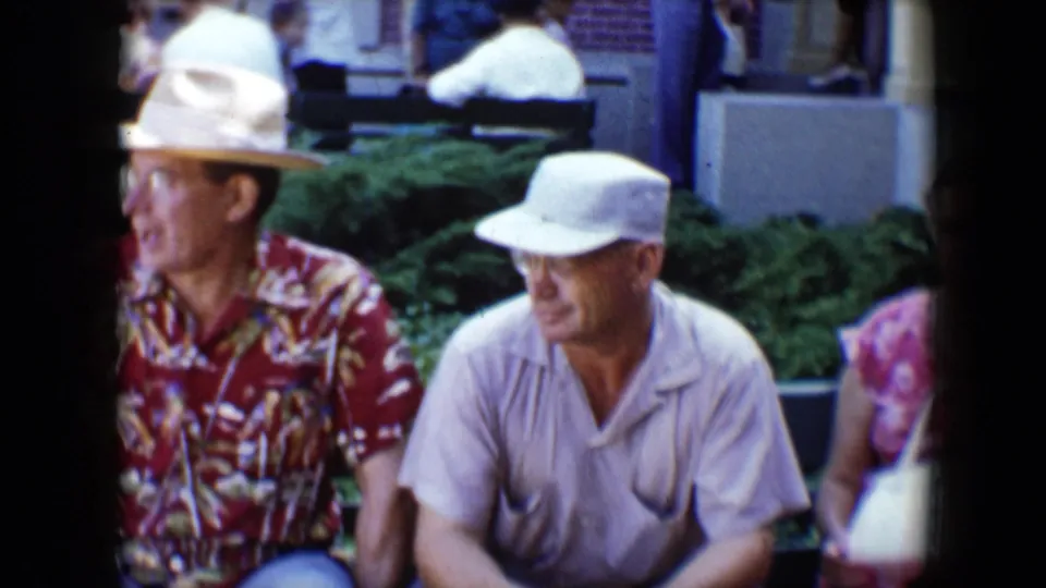 At Disneyland in 1959, a man in a red patterned shirt sits or leans near a man in a white cap, with the woman partly visible at the edge.
