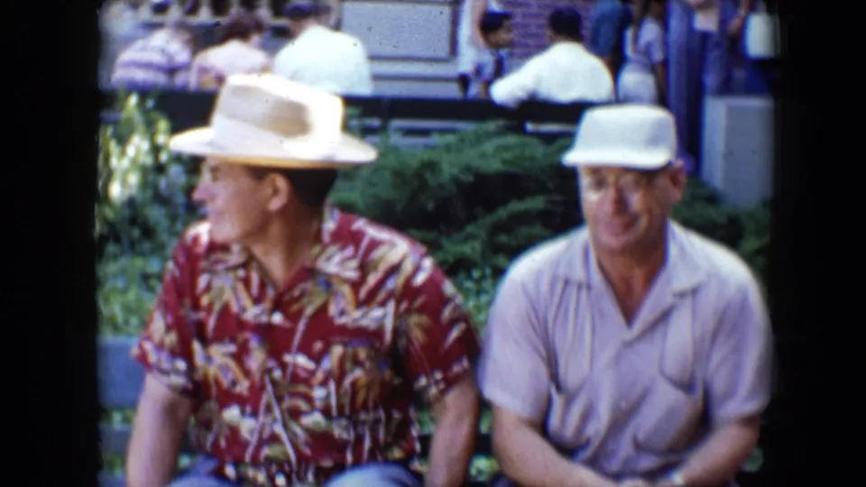 A 1959 Disneyland frame shows two seated men, one in a red patterned shirt and brimmed hat and one in a white cap, with landscaping and visitors behind them.