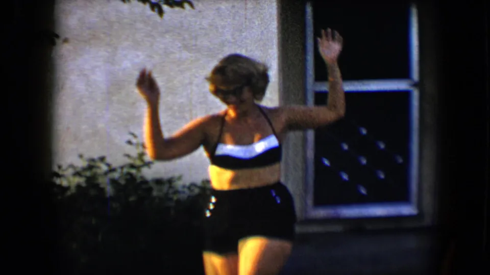 In metadata-supported 1959 home-movie footage, a young woman in a swimsuit stands on a backyard lawn with shrubs and a wall behind her.