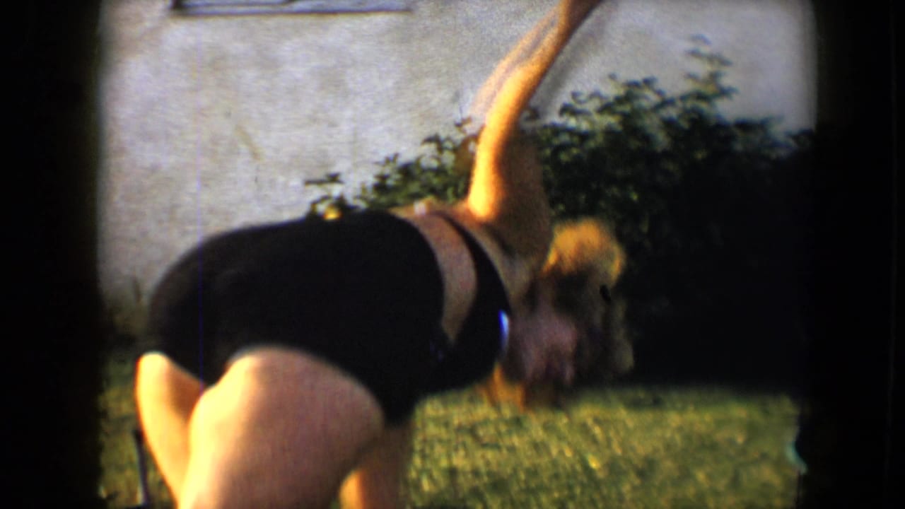 Woman in a swimsuit bends forward mid-cartwheel on grass beside shrubs and a wall.