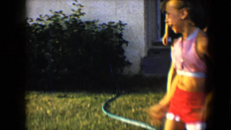 A young woman in a red and white swimsuit stands at the edge of a backyard lawn, with a hose, shrubs, wall, and window visible.