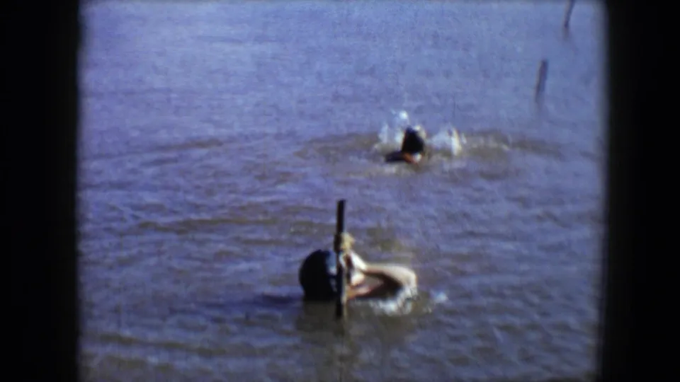 1959 Disneyland home-movie frame showing two swimmers separated by distance in rippling water, with a dark post in front.