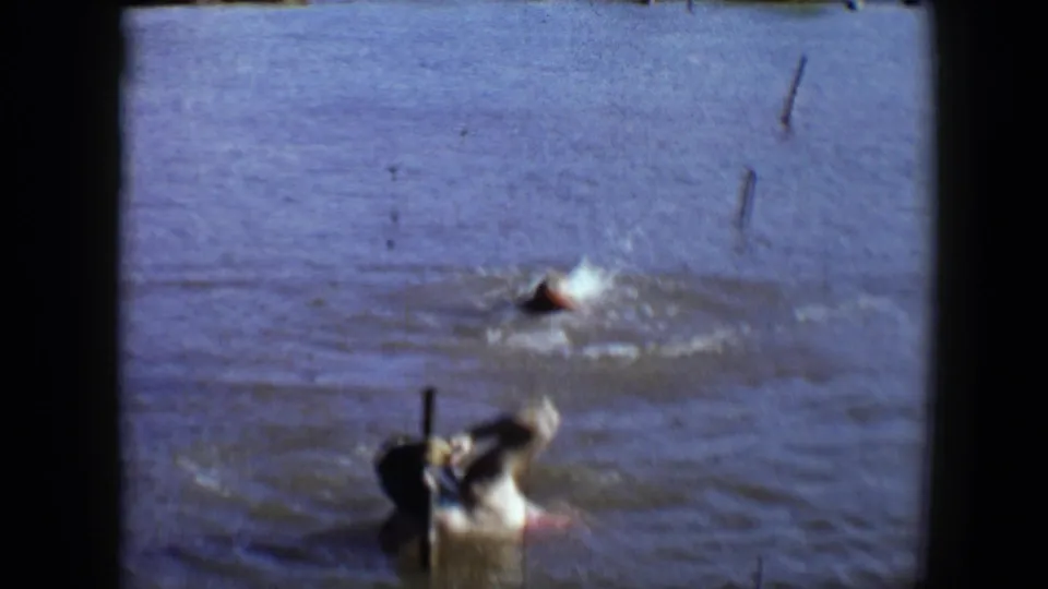 1959 Disneyland frame with a nearer swimmer partly submerged beside a wooden post and a second swimmer splashing farther away.