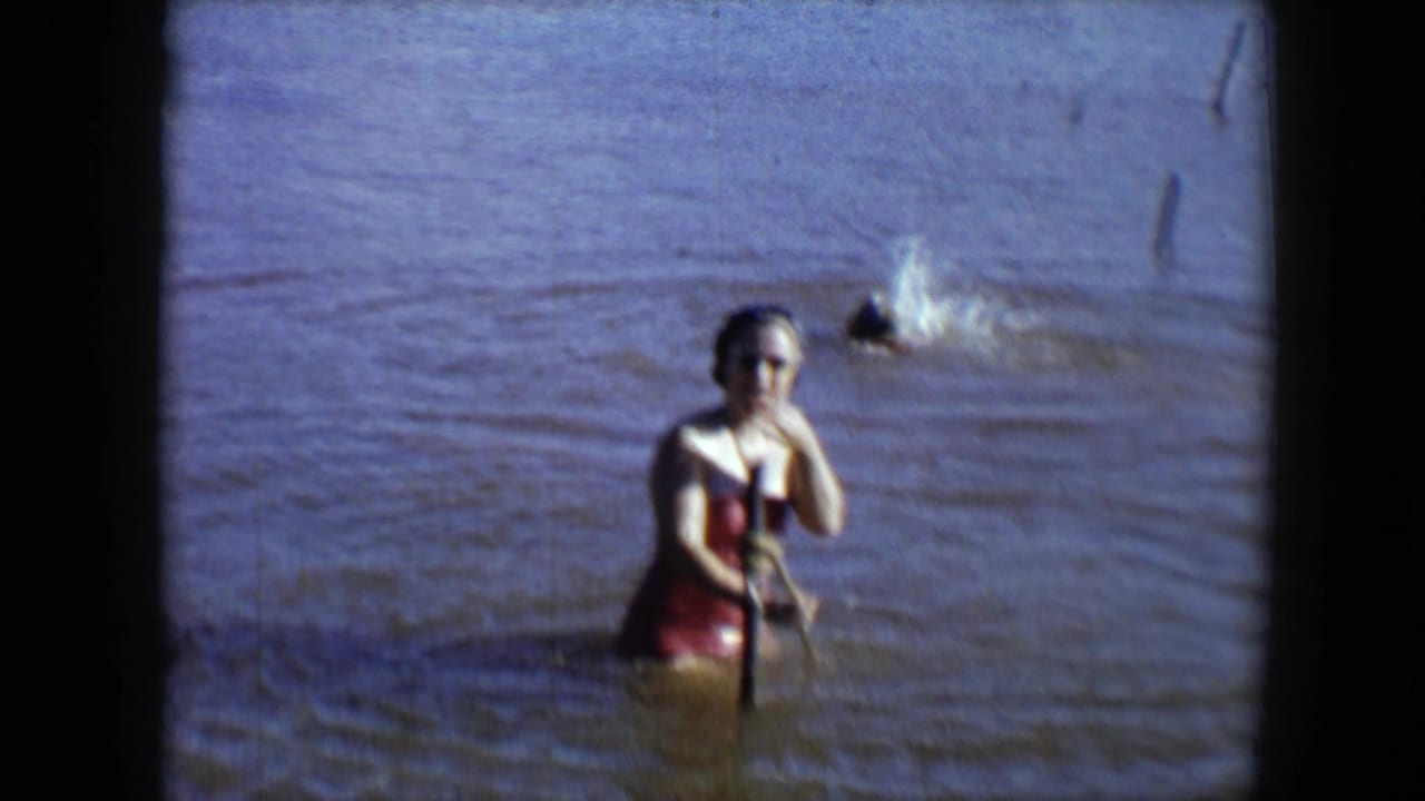 Woman in red swimsuit and sunglasses stands in water near a splashing swimmer.