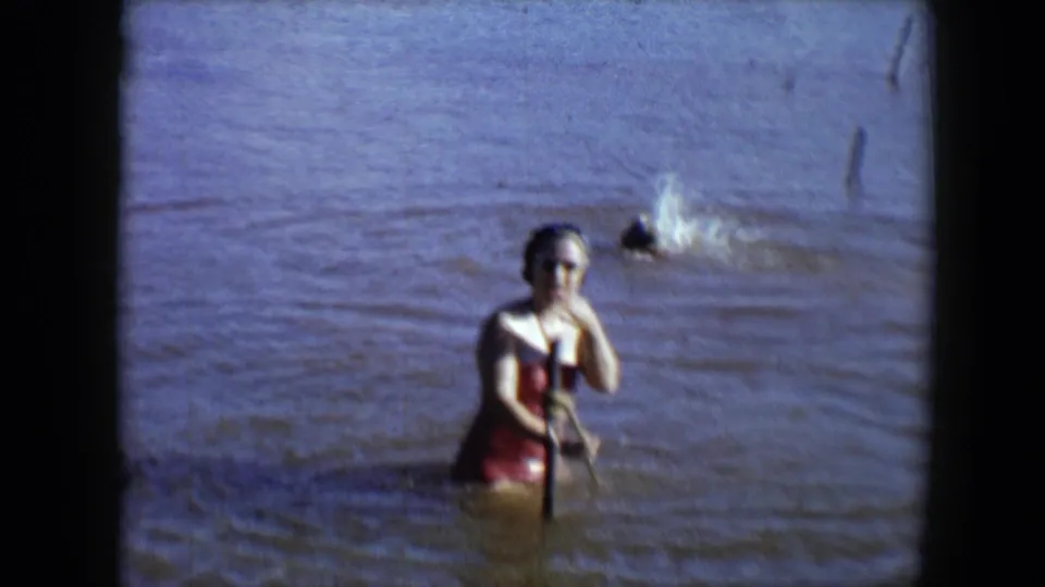 At Disneyland in 1959, a woman in a red swimsuit and sunglasses stands in muddy water near a wooden pole and background swimmer.